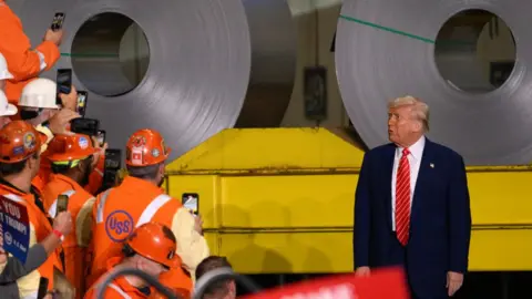 Donald Trump speaking to supporters at a steel factory. He is wearing a dark suit, a white shirt and red tie as men wearing hi-vis jackets and hard hats take pictures of him