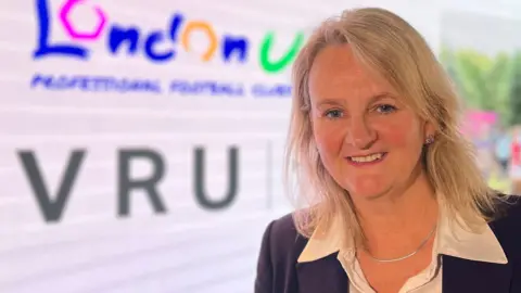 BBC/Harry Low Lib Peck smiles at the camera. She is wearing a white shirt under a navy jacket in front of a VRU sign