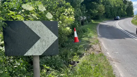 A black sign with an arrow on it on the side of the A36 in Wiltshire. The road next to the sign curves to the right. There are two cars travelling in opposite directions on the road. There is a traffic cone on the grass verge near the sign.