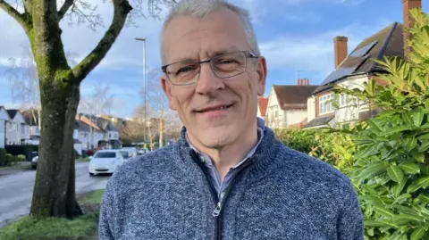 A man wearing a blue jacket with a zip stands outside on a residential road.