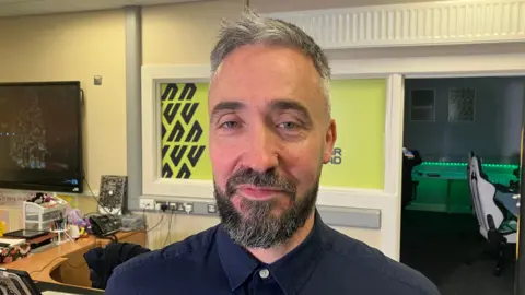 Peter Wilmont smiles. He is wearing a blue shirt with white buttons. He has grey hair and a grey beard. Behind him an open door to the gaming room.