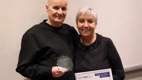 SUPPORT DOGS Janet and Peter are stood together with arms around each other. They're holding a certificate and a small, glass trophy.