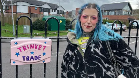 The box is pink and says 'trinket trade' on it. Ashleigh is pictured next to it and has blue hair and is wearing a black jumper. She has pink eye makeup on and has tattoos on her neck. 