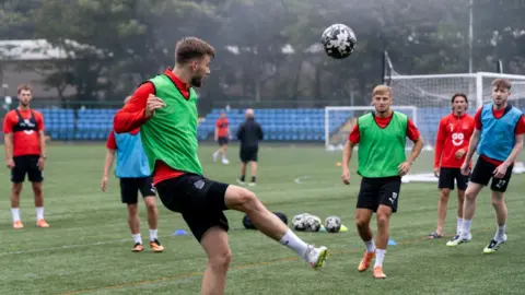 A man kicks a football in the air on a green pitch. Other players in green and blue bibs look on, with two goals in the distance.