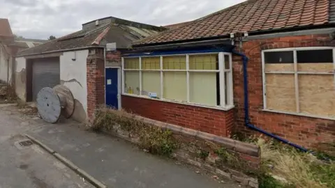 A red brick house with boarded windows and blinds next to a garage with a large wooden cable drum in front of a corrugated iron door