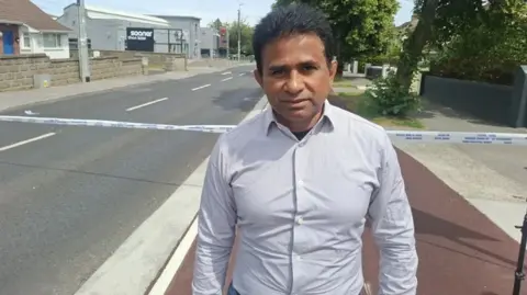 RTÉ Thomas Joseph. He has dark hair, wearing a light coloured shirt. He is standing in front of a police cordon on a residential street.
