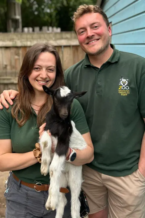Battersea Park Children's Zoo A man with blonde hair and a woman with brown hair smile into the camera, holding a kid goat, which is black and white