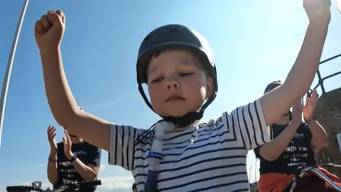 Henry sat on his bike with hands aloft after completing his race he wears a helmet, a ventilator tube and a striped blue and white T-Shirt. behind him other people on bikes are applauding him.