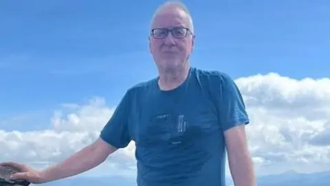 Family handout Gordon Robb, who has short, grey hair and glasses, is wearing a blue T-shirt. He has his left arm outstretched resting on a rock. Behind him the sky is blue with fluffy white clouds.