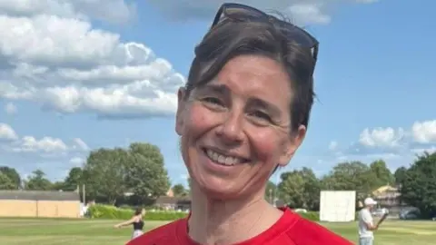Fiona is standing on a green field. She has brown hair in a ponytail, and has sunglasses balanced on top of her head. She is wearing red T-shirt and is holding a trophy and a glass with orange liquid in it.