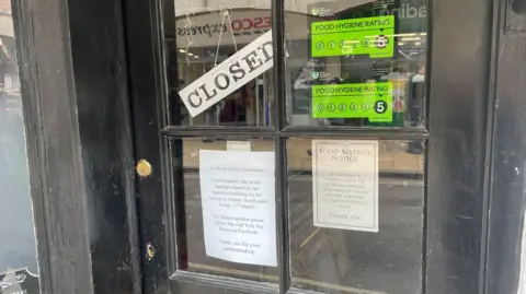 The front door of a cafe. The door is painted black and has several pieces of paper and stickers attached to the glass panels. The scene behind the glass shows part of a street with other shop signs visible in the reflection.