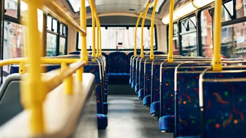 The view towards the back of a public transport bus on the top deck. The rows of seats are dark blue with a colourful swirling pattern, and the handlebars and poles are a bright yellow.