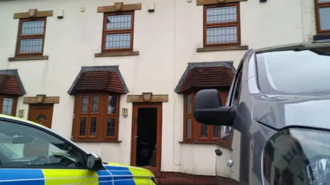 A police car and a black van are parked outside a cream coloured house with brown doors and windows