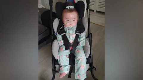 Family handout Lila is pictured in her buggy - which is a dark colour with grey padding for her to sit on. She is strapped into it. She's wearing a matching green floral top and trousers and there's a bow on top of her dark hair. A tube is in her right nostril. The buggy is in a room with a grey wooden floor.