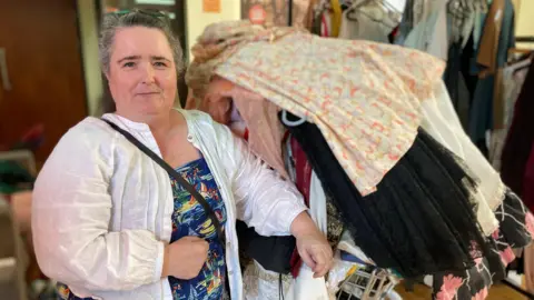 Alex Pope/BBC A woman holding onto a large trolley full of clothes. She has on a dress, a white shirt, and has a bag across her. Her hair is tied back with sunglasses on her head. 