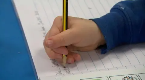 A child wearing a blue sweatshirt is writing in an exercise book with a yellow and black striped pencil.