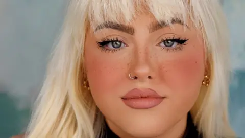 A close-up shot of Megan Lafferty, a young woman with blonde hair and freckled skin wearing glamorous make-up, a black jumper and gold earrings.