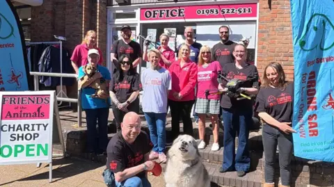 Drone SAR For Lost Dogs IOW A large group of people are pictured gathered outside a building. A man is crouched at the front of the group with a husky. A woman dressed in dark clothing is holding a drone in both hands. A sign to the left reads "Friends of the Animals charity shop" and "open". 