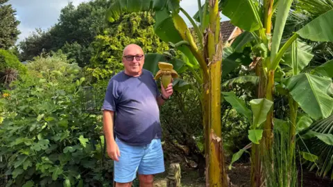 Supplied A head and shoulders image of a man wearing a T-shirt and glasses while holding a banana plant.