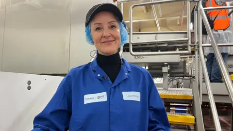 Karl Bird/BBC Manufacturing director Layla Whiting standing in front of machinery wearing a blue overall, cap and hair net.