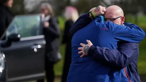 PA Media Two mourners hug each other with a hearse in the background