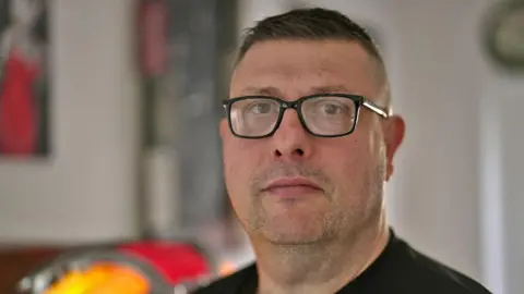 Brian Buckle looks straight towards the camera - he is wearing square glasses and a black jumper. He has short dark hair and dark stubble, and is pictured in a kitchen.