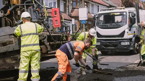 Construction workers laying down tarmac including two holding shovels and one with a large rake-type tool flattening the tarmac. There are lorries in the background and all workers are wearing hi-viz clothing.