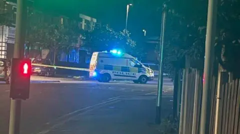 A police van with sirens on is parked in front of a yellow police cordon on a street in the night. There's a pedestrian crossing button at the front of the photo showing a red man light icon.