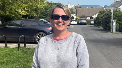 A woman with short blond hair wearing sunglasses and a grey jumper standing in a car park 