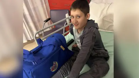 Christina Makin Archie sits on a hospital bed, next to a large overnight bag. He wears grey tracksuit top and bottoms, and has short brown hair.