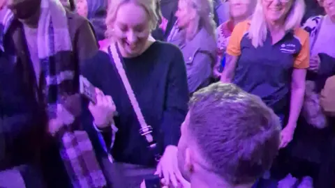 A man on bended knee proposes to a woman at a music concert