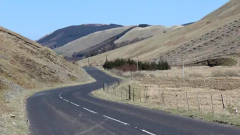 A winding road through the south of Scotland with fencing up one side and rolling hills with a few trees in patches 