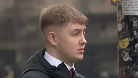 A young man with brown hair, pictured side on, wearing a black coat
