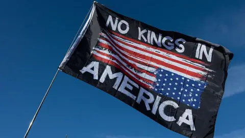 Getty Images A person waves a flag that reads, "NO KINDS IN AMERICA" with the blue sky above it at a Washington DC rally on 17 October - one day before the No Kings protests scheduled in cities across the US. 