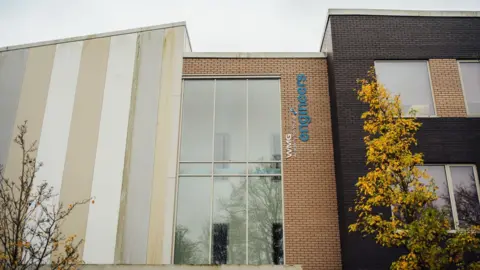 WMG Academy Coventry The exterior of the WMG Academy for Young Engineers building in Coventry. The modern two-storey building has brown brick walls, large glass windows and dark grey cladding, with the school's name and logo displayed in blue lettering. An autumnal tree with yellow leaves stands in front of the entrance.