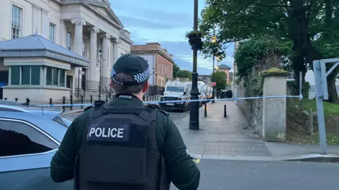A PSNI officer is standing with his back to the camera. He is wearing body armour with the word 'POLICE' written on it. He is looking towards a line of PSNI Land Rovers.
