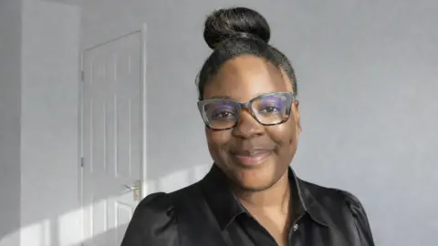 Faith Brynel Pomell A woman with black hair tied up in a bun wearing glasses and a black satin blouse smiling. 