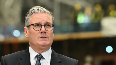 Keir Starmer has short grey hair and is wearing black glasses, a black suit jacket, white shirt and black and white spotted tie