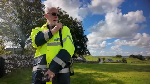 An older man with white hair and dark glasses, he is wearing hi-viz and holding a radio handset. He us standing in a green field in front of a wall and some trees. Behind is a cloudy, blue sky.