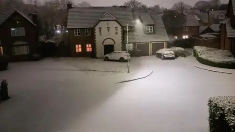 A residential street with houses on and cars parked outside them, all covered in snow. The road is also covered in snow.