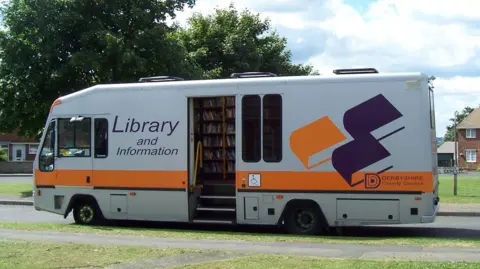 A view of an old Derbyshire County Council mobile library, a 2013 DAF LF45, parked next to a grass verge.