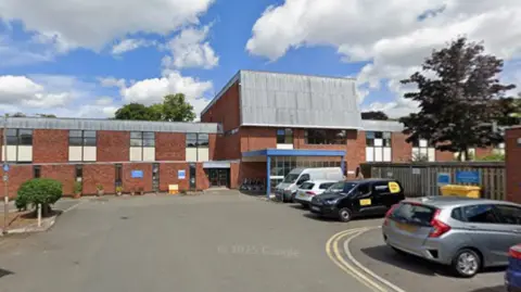 Google A car park in front of a hospital building, with two vans and two cars parked.