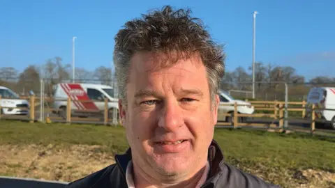 Luke Deal/BBC James Blyth smiles at the camera as he stands outside. He has dark hair which is slightly curled with some grey hair at the sides of his head. He wears a dark jacket with a lighter shirt underneath. White vans can be seen parked behind him on a road. 
