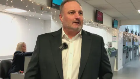 A man with short hair, wearing a black suit jacket and a white shirt, stood in front of an airport check in desk.