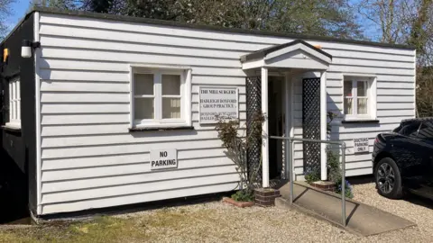 Steve Martin/BBC The single-storey surgery building pictured from further back. A gravel drive leads to its entrance, which has a black trellis and plants either side of the door. The building's sides are black and the front is made of white weatherboards. A small ramp allows for wheelchair access to the door.