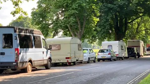 A line of white vans and caravans, some with cars in front or behind, parked along a section of road along The Downs in Bristol