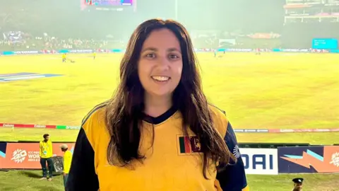 Yasmin stands at a sports game, in a yellow sports top, with a wide smile. The pitch can be seen in the background