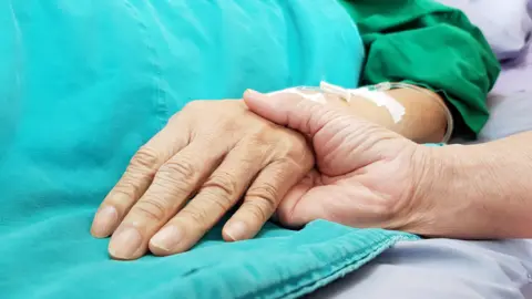 Getty Images The hand of a patient in a hospital bed being held by another, above a blue blanket.