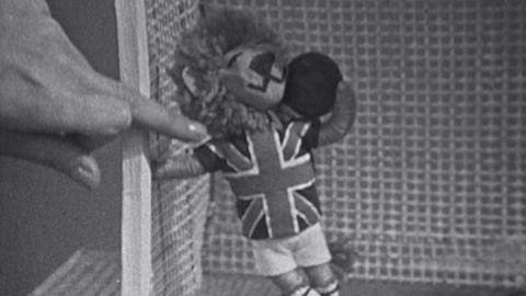Valerie's hand pointing at a small lion doll wearing a Union Jack shirt and holding a football.