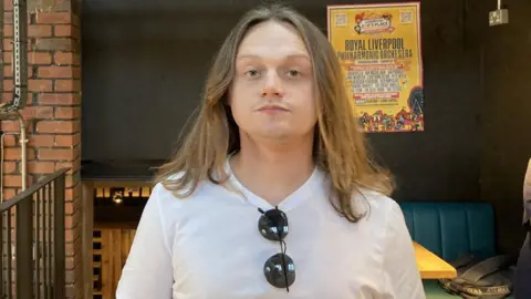 Tom Walker/BBC Jamie Webster has long hair, a white T-shirt with sunglasses hanging on the neck